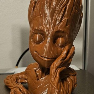 May include: A brown 3D printed figurine of a character from the Guardians of the Galaxy movie, Groot. The figurine is in a sitting pose with one hand resting on its chin. The figurine has a detailed, textured surface that resembles wood.