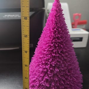 Puede incluir: Un vibrante árbol de Navidad decorativo magenta, de aproximadamente 15 cm de altura. El árbol tiene un aspecto texturizado y erizado, con forma cónica. El árbol se encuentra sobre un fondo borroso, con una cinta métrica en el marco.