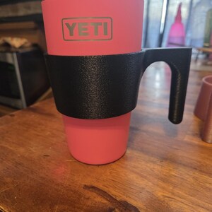 May include: A pink Yeti tumbler with a black handle and a clear lid. The Yeti logo is visible on the front of the tumbler. The tumbler is sitting on a wooden surface.