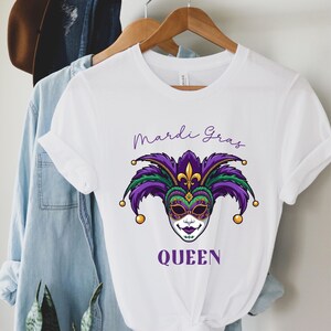 以下が含まれることがあります： 紫、緑、金、黒のマルディグラマスクのデザインが施された白いTシャツ。「Mardi Gras」の文字がマスクの上に、「QUEEN」が下に、すべて紫で書かれています。