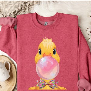Puede incluir: Una sudadera roja con un dibujo de un pato de dibujos animados soplando una burbuja rosa. El pato tiene plumas amarillas, ojos azules y una pajarita floral. La sudadera tiene mangas largas y cuello redondo.