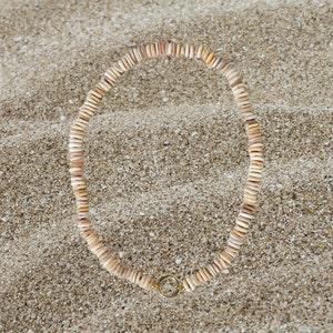 Könnte beinhalten: Eine Halskette aus kleinen, flachen, hellfarbigen Muscheln, die kreisförmig angeordnet sind. Sie wird mit einem goldfarbenen Verschluss geschlossen. Der Schmuck liegt auf sandigem Untergrund und erinnert an ein Strand- oder Küstenmotiv.