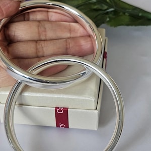 May include: Two polished silver bangles, reflecting light, are interlocked. The smooth, circular bands are presented against a neutral backdrop, hinting at jewellery or accessories. The bangles appear to be made of metal, possibly sterling silver.