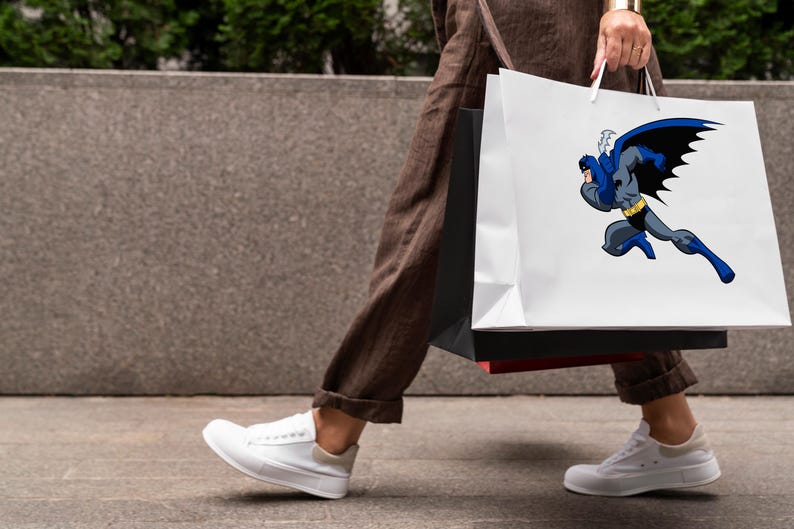K&ouml;nnte beinhalten: Eine Person tr&auml;gt zwei Einkaufstaschen. Die Tasche rechts hat einen wei&szlig;en Hintergrund mit einem Comicbild von Batman in Blau und Schwarz. Die Tasche links ist schwarz.