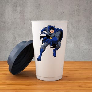 K&ouml;nnte beinhalten: Eine wei&szlig;e Reise-Tasse mit einem blauen und grauen Cartoon-Bild von Batman auf der Vorderseite. Die Tasse hat einen dunkelblauen Deckel.
