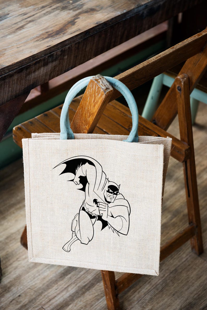 K&ouml;nnte beinhalten: Eine beige Jute-Tragetasche mit einem blauen Griff. Die Tasche zeigt eine Schwarzwei&szlig;-Illustration von Batman in einer Superheldenpose.