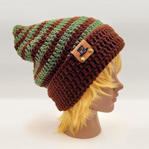 Puede incluir: Un gorro de ganchillo hecho a mano en tonos marrones y verdes. El sombrero presenta rayas alternas marrones y verdes, con un borde marrón liso. Una pequeña etiqueta de cuero con un logotipo está adherida al borde.