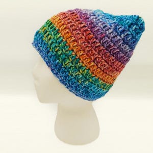 May include: A handmade, rainbow-colored crochet beanie hat. The hat is displayed on a white mannequin head. The hat features a vibrant spectrum of colors, including blue, green, yellow, orange, and purple. The beanie is a stylish accessory.