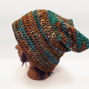 May include: A slouchy beanie hat crafted from thick, variegated yarn in earthy tones of brown, tan, and deep teal. The crochet construction features a textured, ribbed stitch pattern that creates a soft, flexible shape with a rounded top.