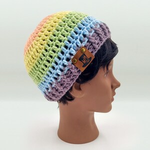 May include: A crochet beanie featuring a horizontal rainbow gradient pattern in pastel shades of peach, yellow, green, blue, and purple. The hat includes a brown rectangular leather tag on the side with a debossed top hat icon and the letters MH.