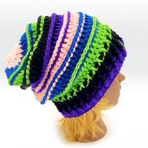 May include: A colorful, handmade crochet beanie in a slouchy style. The hat features a vibrant mix of colors, including pink, blue, green, black, and purple. The yarn is arranged in vertical stripes, creating a textured look.