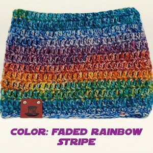 May include: A crocheted cowl in a faded rainbow stripe pattern. The cowl features a spectrum of colors, including blue, purple, orange, and green. A small brown leather tag with a logo is attached to the cowl. The text "COLOR: FADED RAINBOW STRIPE" is below the cowl.