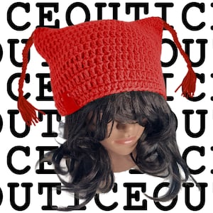 Op de afbeelding: Een felrode gehaakte muts met puntige oren en kwastjes. De muts wordt gedragen op een mannequinhoofd met donker haar. De woorden "ICE OUT" zijn in een herhalend zwart-wit patroon rond de muts gedrukt.