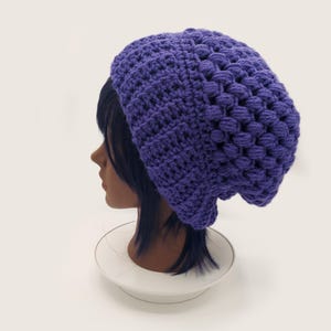 Könnte beinhalten: Eine leuchtend violette Häkelmütze mit einem strukturierten Noppenmuster an der Krone und einem gerippten Band am Saum. Die Mütze wird auf einem Schaufensterpuppenkopf präsentiert und zeigt die lockere Passform sowie die komplexe Garnarbeit.