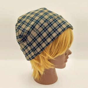 May include: A plaid beanie in shades of tan, navy blue, and white. The hat is worn on a mannequin head with yellow hair. The beanie has a classic plaid pattern, offering a stylish accessory for various outfits.