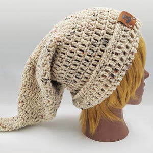 Peut inclure: Chapeau en crochet crème avec une longue extrémité pointue. Le chapeau a un aspect texturé avec des mouchetures marron et orange. Une petite étiquette en cuir marron est attachée sur le côté. Un accessoire unique.