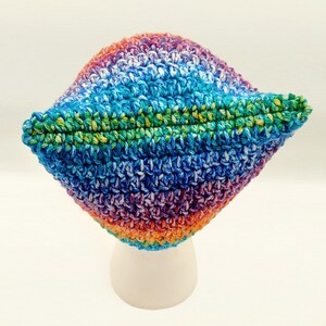 May include: A colorful, handmade crochet hat. The hat features a vibrant mix of colors, including blue, green, purple, orange, and white. The hat is displayed on a white stand against a white background. The hat's brim is folded up.