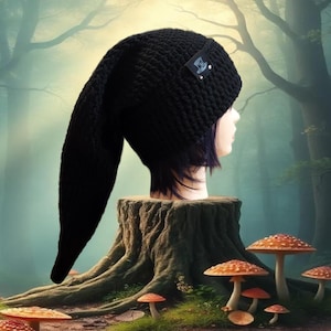 May include: A black, handmade crochet hat with a long, pointed tip. The hat is displayed on a mannequin head. A small tag with a logo is attached to the hat. The background features a forest scene with mushrooms.