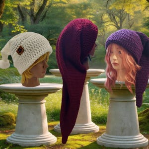 May include: Three crochet hats displayed on mannequin heads. One is cream-colored with a pom-pom and a small brown tag, one is a long deep burgundy pointed cap, and one is a long purple pointed cap worn over wavy pink hair.