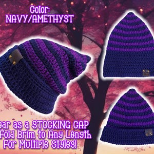 May include: A navy blue and amethyst striped crocheted beanie hat with a fold-over brim. The hat can be worn as a stocking cap or with the brim folded to create different styles. "Color: NAVY/AMETHYST" is written on the image.