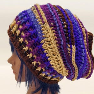 May include: A handmade crochet beanie in a mix of colors, including brown, tan, purple, and blue. The hat has a textured, striped design and is shown on a mannequin head. This winter hat is a cozy accessory.