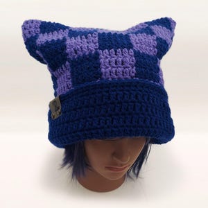 May include: A handmade crochet hat in a cat ear design. The hat features a checkerboard pattern in shades of purple and blue on the ears, with a solid blue brim. A small, rectangular tag is attached to the brim.