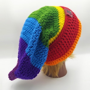 Könnte beinhalten: Ein handgefertigter Häkelhut in Regenbogenfarben, darunter Rot, Orange, Gelb, Grün, Blau und Lila. Der Hut hat ein langes, lockeres Design und ist mit einem strukturierten Stich gefertigt. Ein kleines Lederetikett ist an der Seite befestigt.