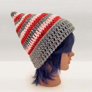 May include: A crocheted beanie with a pointed top, featuring horizontal stripes in alternating colors of grey, red, and light pink. The hat has a wide, textured grey brim worked in a vertical stitch pattern that contrasts with the main body.