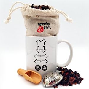 May include: A white mug with black arrows and the letters "B" and "A" sits next to a wooden scoop and a silver tea infuser. A small burlap bag with the text "apple & salt" embroidered in red holds loose tea leaves, and is placed on top of the mug.
