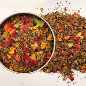 May include: A metal tin filled with a colorful blend of dried herbs and flowers, including red rose petals, orange pieces, and lavender. A pile of the same mixture is scattered beside the tin, creating a visually appealing arrangement.