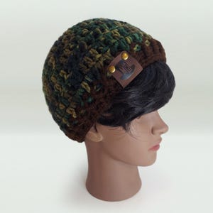 May include: A crocheted beanie featuring a camouflage pattern of dark green, olive, and brown yarn. The hat has a solid brown ribbed brim and a square brown leather patch attached with two gold-colored metal rivets, displaying a top hat graphic.