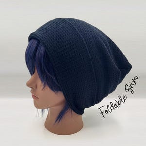 May include: A dark navy blue beanie with a textured knit pattern and a foldable brim. The beanie is displayed on a mannequin head with purple hair. The text "Foldable Brim" is printed in black script on the right side of the image.