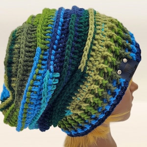 May include: A handmade crochet hat in shades of blue and green. The hat features a textured stitch pattern and a slouchy fit. A small, dark blue label with silver rivets is attached to the side. This hat is a stylish accessory for cold weather.