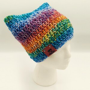 May include: A handmade, colorful crochet hat in a rainbow of hues, including blue, green, yellow, orange, and purple. The hat has a cat ear design and a brown leather tag with a logo. This cozy hat is perfect for adding a pop of color to any outfit.