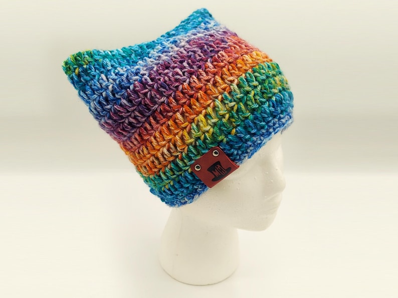 May include: A handmade, colorful crochet hat in a rainbow of hues, including blue, green, yellow, orange, and purple. The hat has a cat ear design and a brown leather tag with a logo. This cozy hat is perfect for adding a pop of color to any outfit.