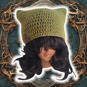 Puede incluir: Un gorro de punto de color verde con un parche de cuero. El gorro lo lleva una persona con el pelo largo y oscuro.