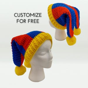 May include: Two crocheted hats with alternating red, blue, and yellow vertical stripes and a yellow pom-pom at the tip. The hats feature a yellow ribbed brim. The text CUSTOMIZE FOR FREE is visible in the image. The hats are displayed on white heads.