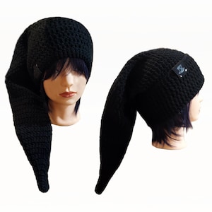 May include: Black crochet hat with a long, pointed extension. The hat is displayed on a mannequin head, showcasing its unique design. A small label is attached to the hat. The hat is made of a thick, textured yarn.
