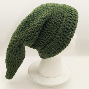 Può includere: Un cappello all'uncinetto verde oliva fatto a mano con una lunga patta a punta. Il cappello è esposto su una testa di manichino bianca. La trama del cappello è visibile, mostrando il dettagliato lavoro all'uncinetto. Un accessorio unico.