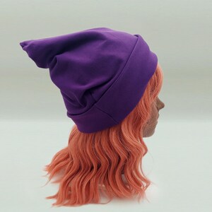 May include: A solid purple beanie hat with a slouchy, pointed back design and a wide, folded cuff at the base. The hat is made of a smooth, matte fabric and features a simple, clean construction with visible seams along the sides and top.