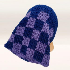 May include: A blue and purple crochet hat with a checkerboard pattern. The hat is handmade and features a brim that extends down the back. The colors are a deep blue and a lighter purple, creating a striking contrast. The hat is likely for cold weather.