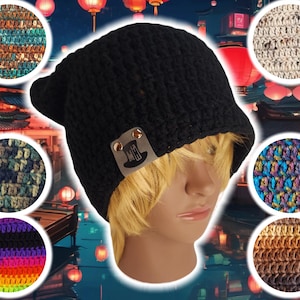 以下が含まれることがあります： 「The Hat Guy」と書かれた小さな革のパッチが付いた黒いクロシェのビーニー。ビーニーはブロンドの髪の人の頭にかぶっています。背景は赤い提灯のぼやけた画像です。