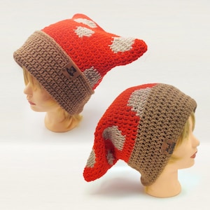 May include: Two crochet beanies featuring a bright red top with beige spots and a solid brown ribbed brim. Each hat includes a small rectangular brown leather-like tag attached to the side, displaying a graphic of a top hat and text.