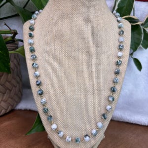 Puede incluir: Collar de cuentas redondas y moteadas en tonos verdes, blancos y grises, ensartadas en una cadena dorada. Las cuentas tienen aproximadamente 1,3 cm de diámetro. El collar se exhibe en un maniquí, resaltando su diseño y colores.