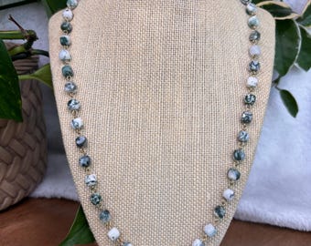 May include: A beaded necklace displayed on a mannequin. The necklace features round, speckled beads in shades of green, white, and gray, strung on a gold-colored chain. The beads are approximately 0.5 inches in diameter. The necklace is presented against a neutral-toned background.