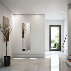 Può includere: Un corridoio moderno con uno specchio a figura intera, un'opera d'arte incorniciata e una pianta in vaso. Lo specchio ha una cornice nera e riflette il corridoio. Le pareti sono bianche e il pavimento è piastrellato. Una porta alla fine del corridoio conduce a un'area esterna.