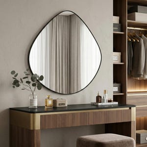 Puede incluir: Un tocador moderno con un espejo con marco negro de forma irregular. El tocador tiene una encimera oscura, detalles dorados y una base de madera. Los accesorios incluyen frascos de perfume, una bandeja de mármol y un jarrón con vegetación.