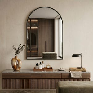 May include: A contemporary dressing table setup with a large arched mirror in a black frame. The wooden vanity has a marble top, a decorative vase, and a tray with perfume bottles. A black lamp and books are also on the vanity.