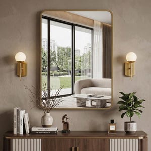 May include: A rectangular gold-framed mirror hangs above a wooden cabinet. Two gold sconces with spherical lights flank the mirror. The cabinet displays books, a vase with branches, a sculpture, a perfume bottle, and a potted plant.
