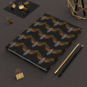 May include: A black notebook with a repeating pattern of a death's-head hawkmoth, a type of moth with a skull-like pattern on its thorax. The notebook is lying on a dark brown surface with two pencils beside it.